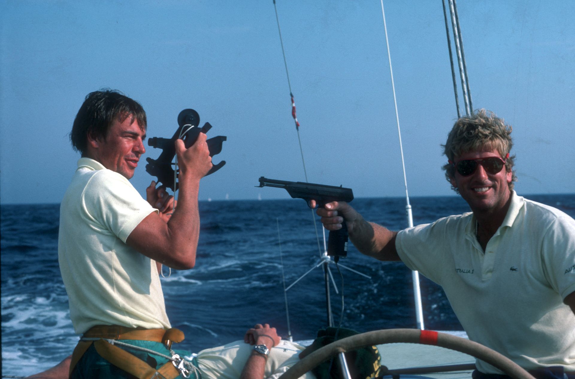 On the deck of the yacht, two crew hold a pistol-shaped tool and a sextant with grins on their faces. Behind them a third man relaxes, lying on the deck.