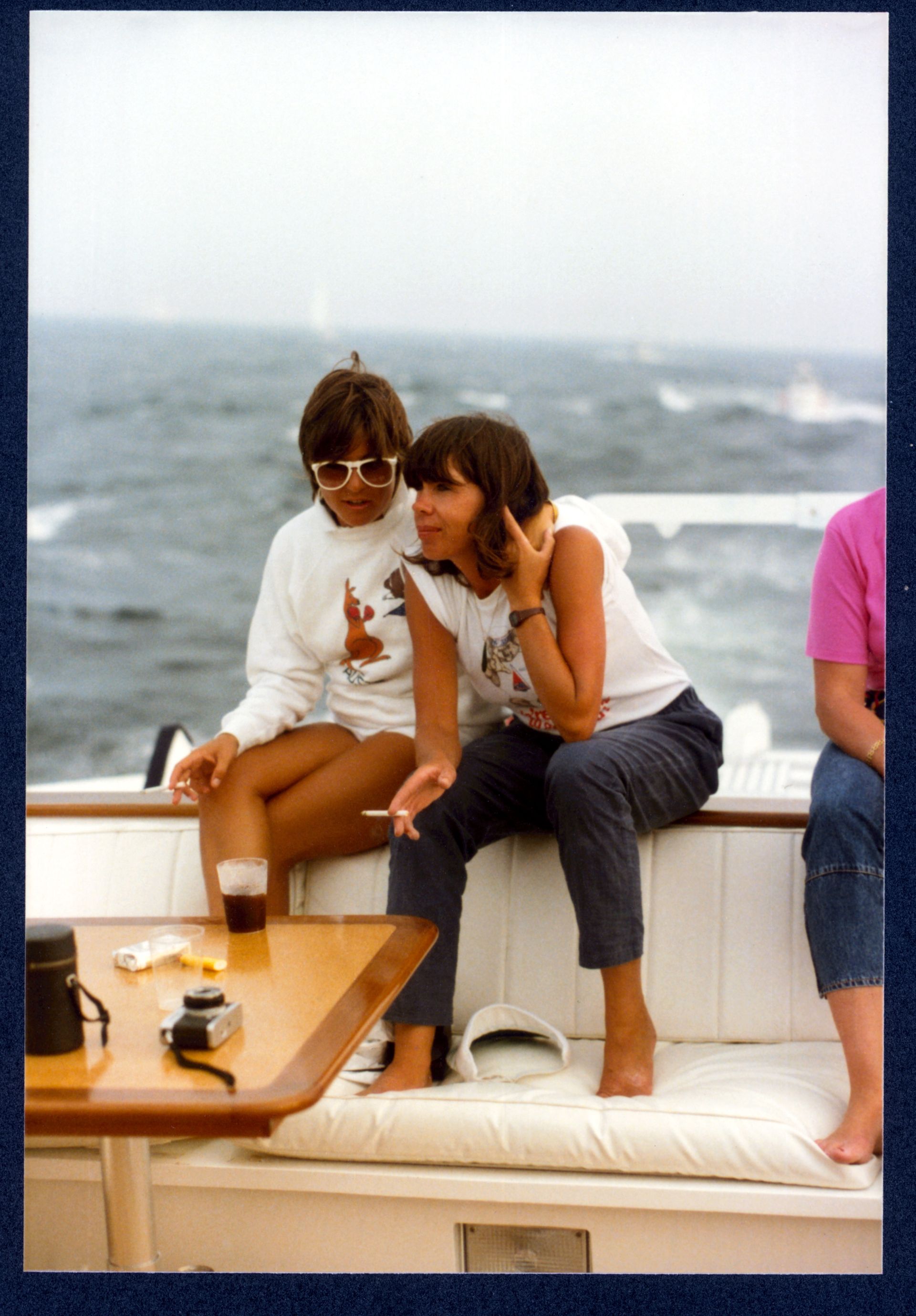 Two young women in 1980s clothing, both with cigarettes, are perched at the edge of a boat's deck. One has an arm around the other as they chat.