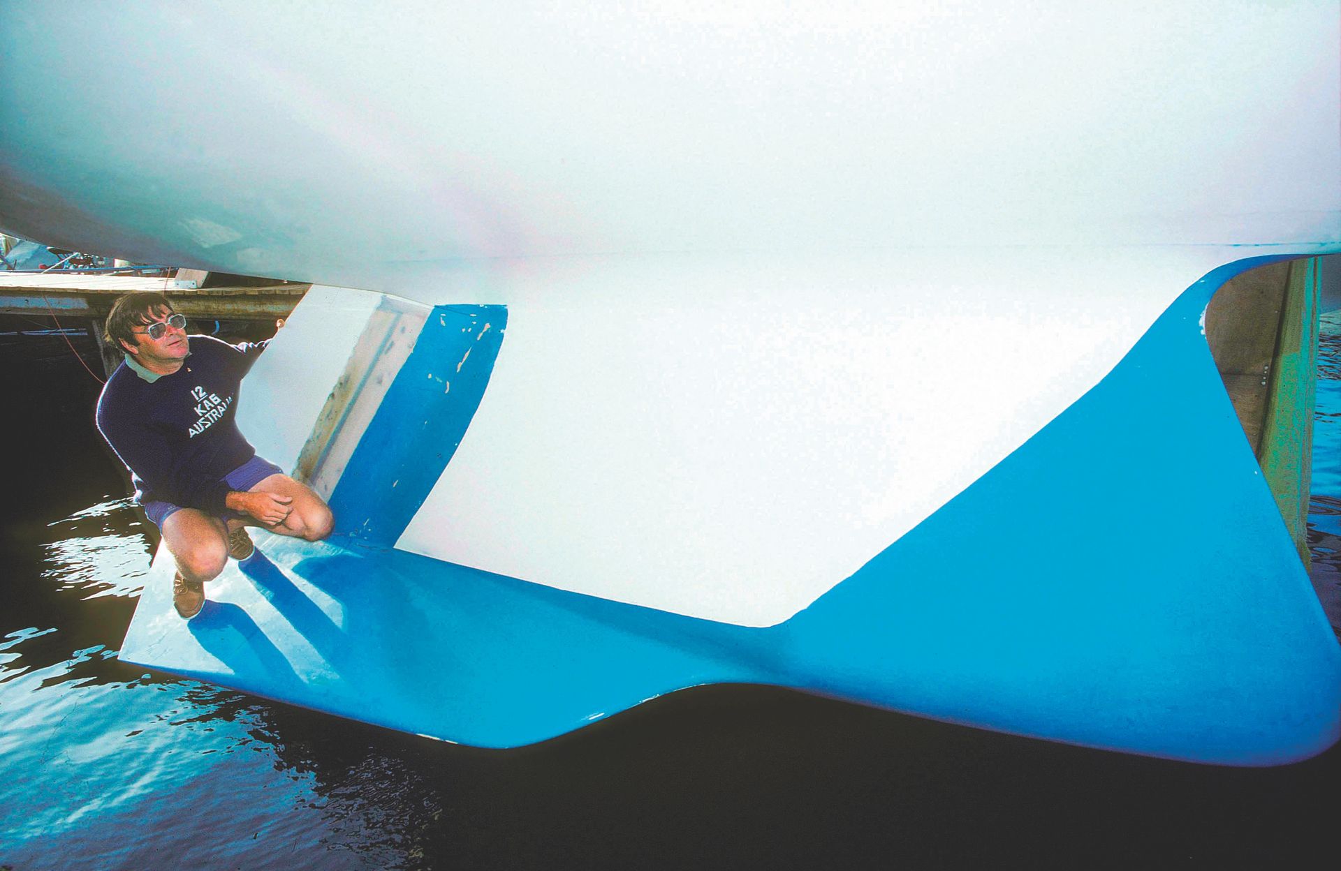 The blue and white keel is suspended above the water's surface. Ben perches on the keel's side, gripping the back of it to avoid falling in the water.