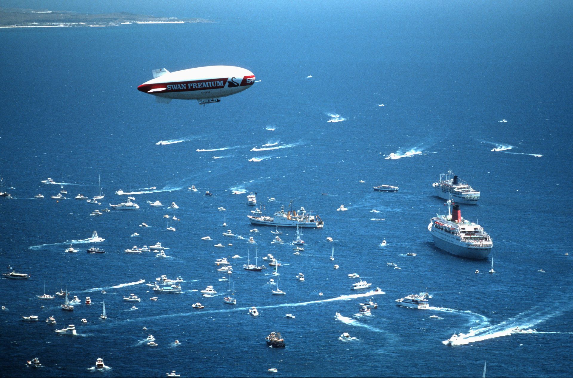 A blimp with the logo "Swan Premium" hovers above a large group of vessels on the water, from small boats to large ships.