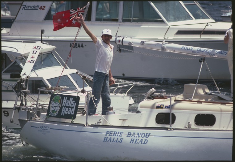Jon Sanders waving from the back of a yacht in harbour, in front of an Australian flag.