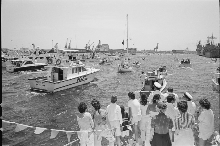 Onlookers crowd the bow of a boat in the foreground, looking at Jon Sanders' yacht in the centre of the photo. A Police boat and many other boats are also sailing with Jon Sanders.