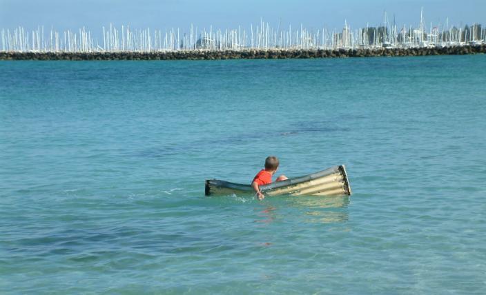A boy in a red shirt paddles a tiny tin canoe over the expanse of the Fremantle Harbour.