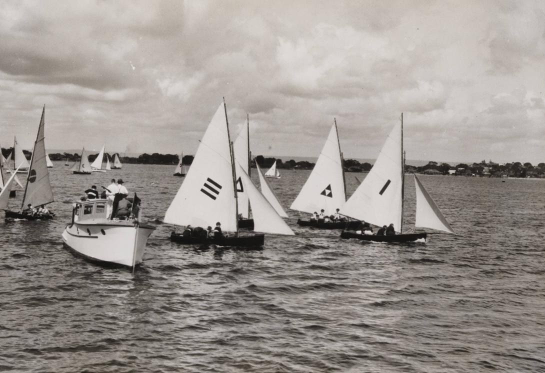 A black and white photo of many small yachts sailing. A motor boat is nearby with people standing observing the race.
