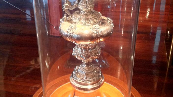A large ornate silver trophy cup behind curved glass.