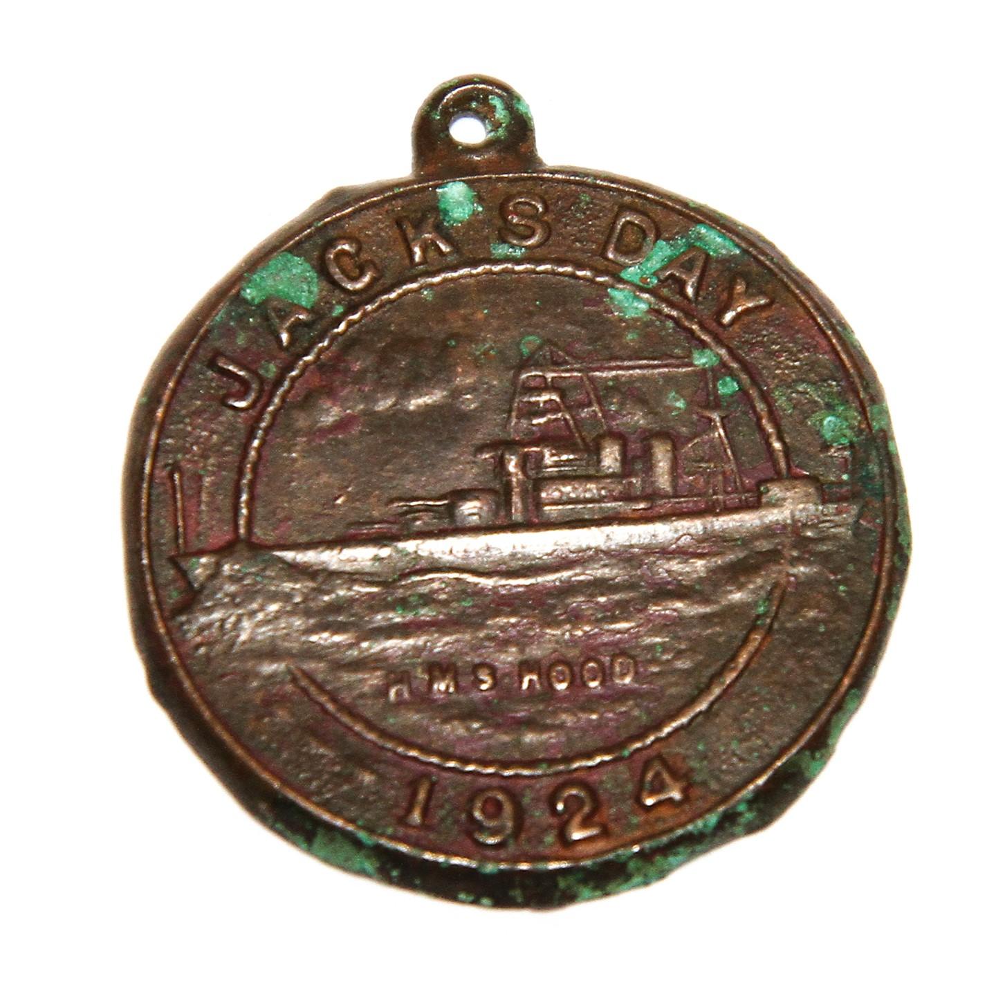 A brown metal medal, with green corrosion spots. There is an image of a ship in the centre and the medal reads 'Jacks Day, HMS Hood, 1924.'