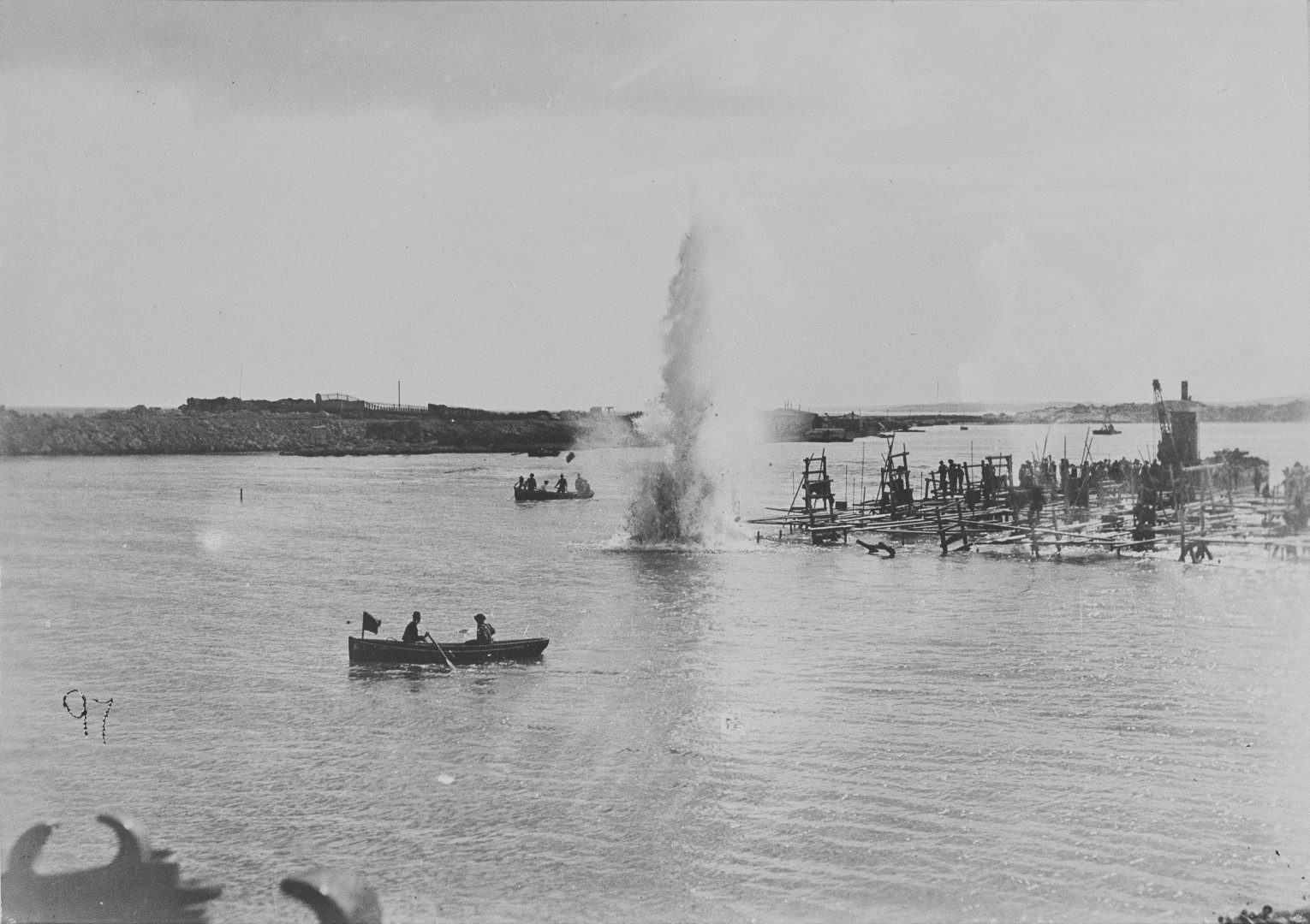 A photograph of an explosion in the harbour. There are workers watching from boats and a makeshift platform.