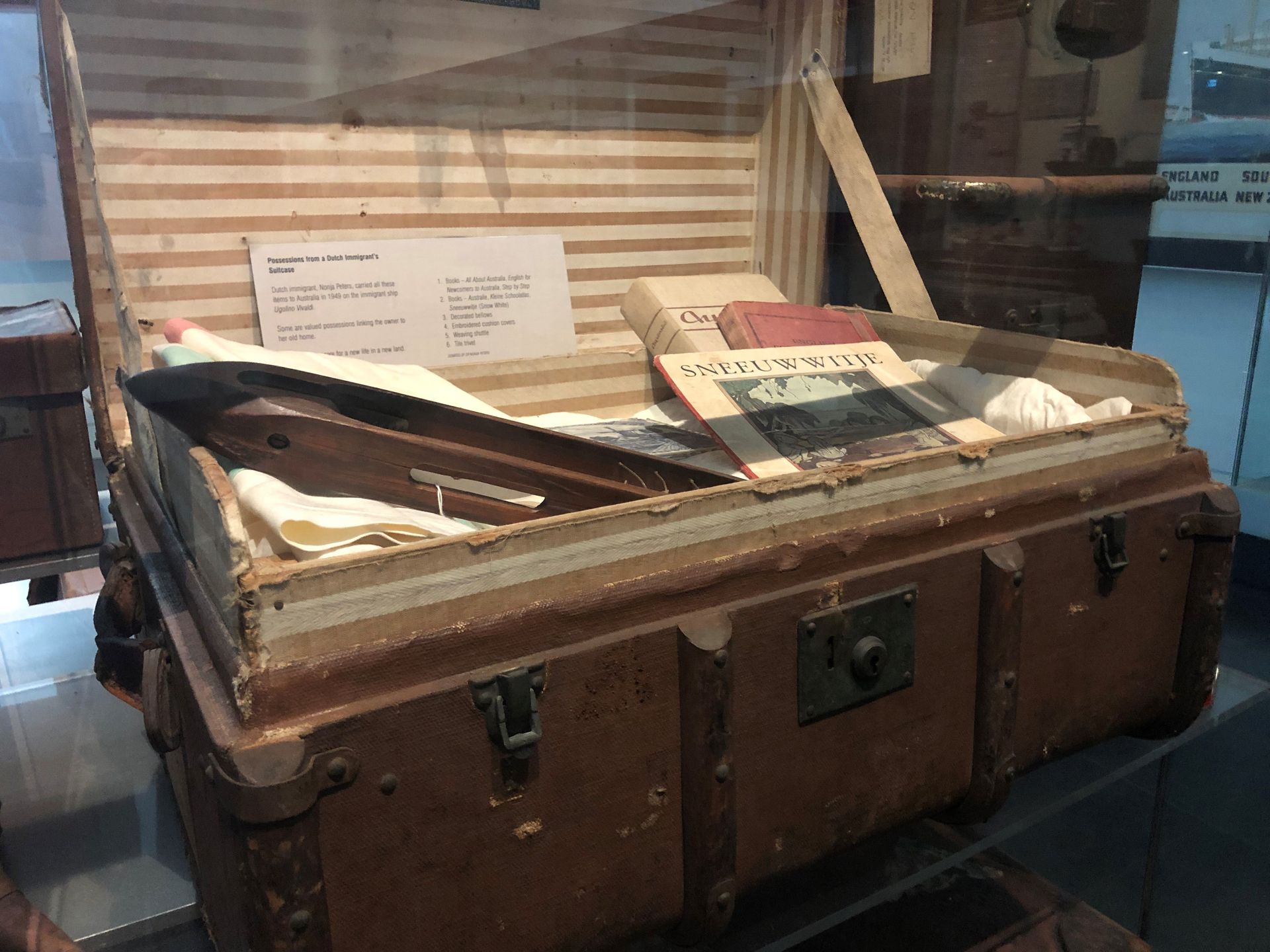 Inside a display case is a very worn old suitcase. It is opened to show items of clothing, and children's books written in Dutch.
