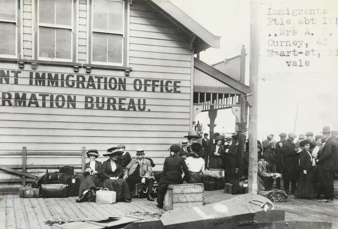 Men and women wearing early 1900s clothing rest next to the Immigration Office. They have their luggage on the ground beside them.