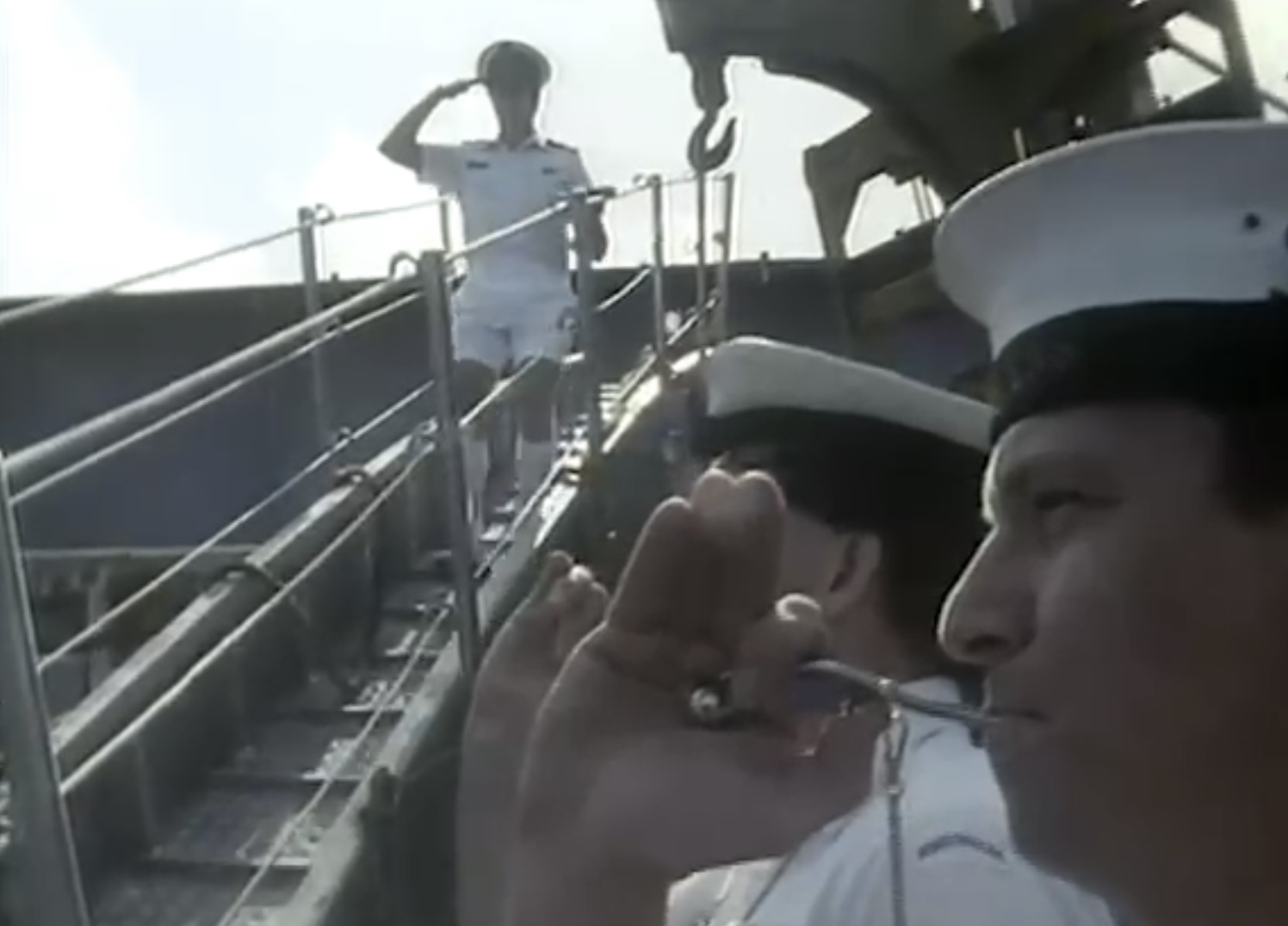 Two sailors blowing small musical instruments stand beside a walkway as a man in a white uniform and hat walks down it, saluting.