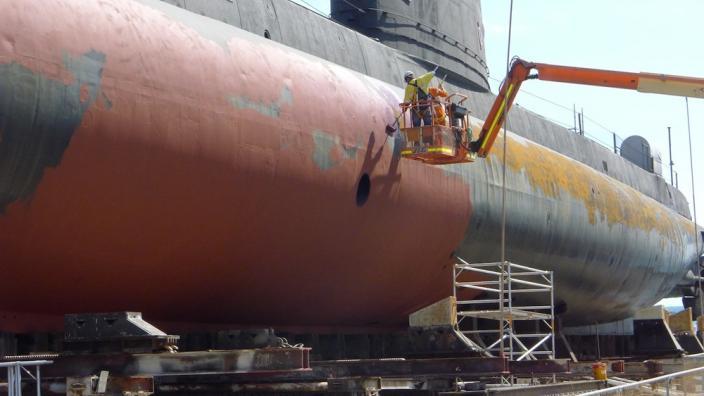 A worker on a crane platform is lifted up to the submarine's side. Part of the submarine's paint is scraped away.