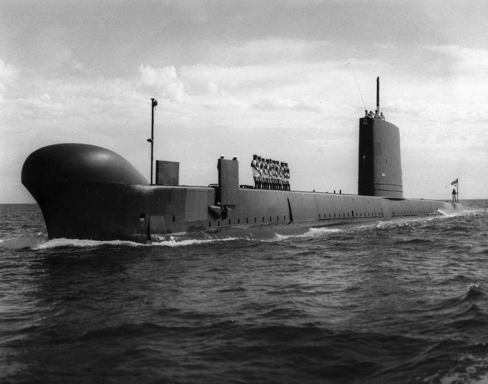 Members of the Royal Australian Navy in clean white uniforms stand on the submarine's deck. Several others are on the tower and one is at the stern with an Australian flag.
