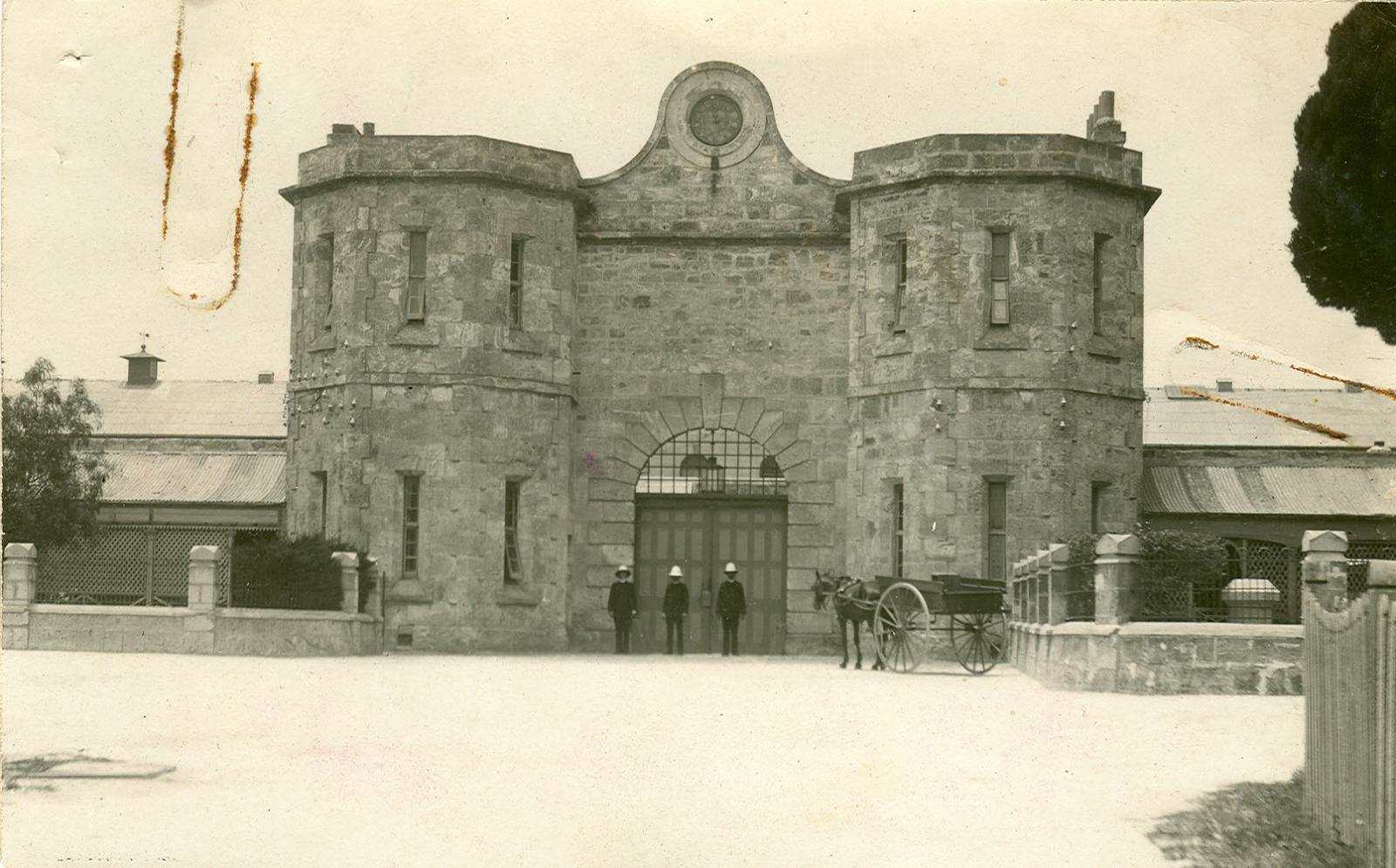 A large stone gate house with two turrets. Three soldiers in white helmets stand at attention and a horse and cart is beside them.