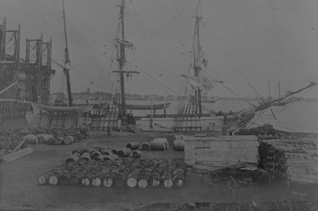 A three-masted ship at dock. A cart and many barrels are on the dock.