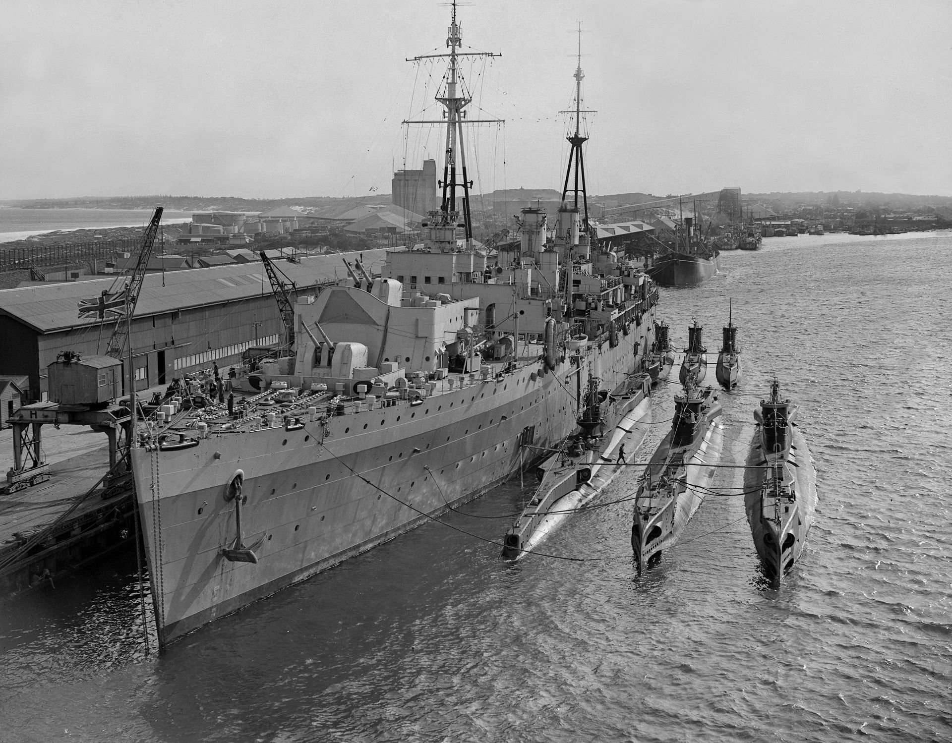 A warship flying a British flag docked at the harbour. Beside it two rows of three submarines each are also docked.