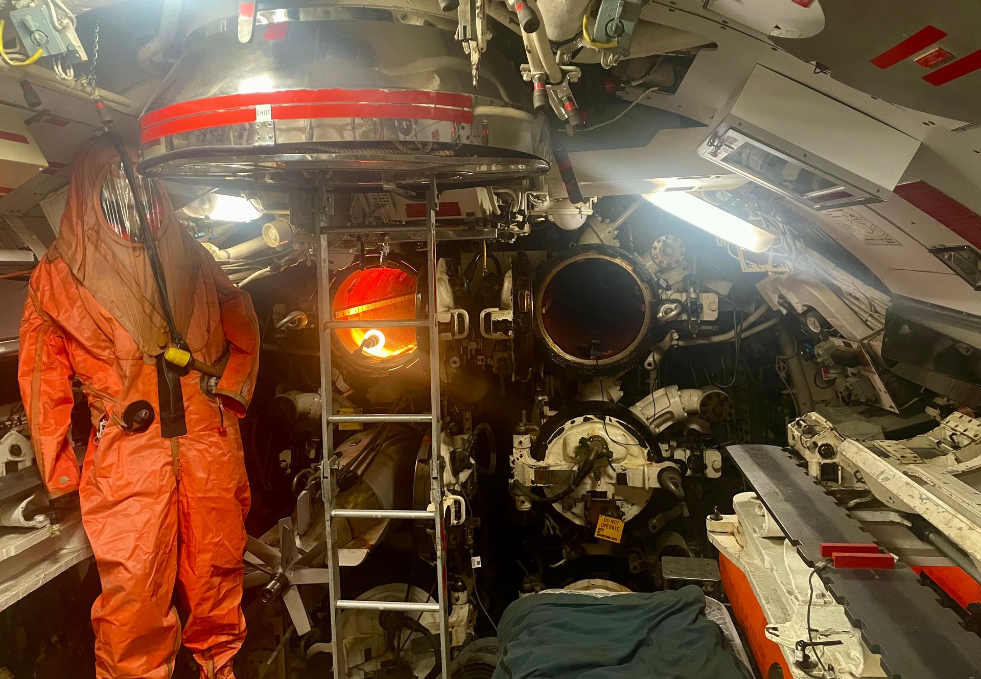 A cramped submarine compartment with four torpedo tubes, and an orange protective suit with face shield hanging from the ceiling.