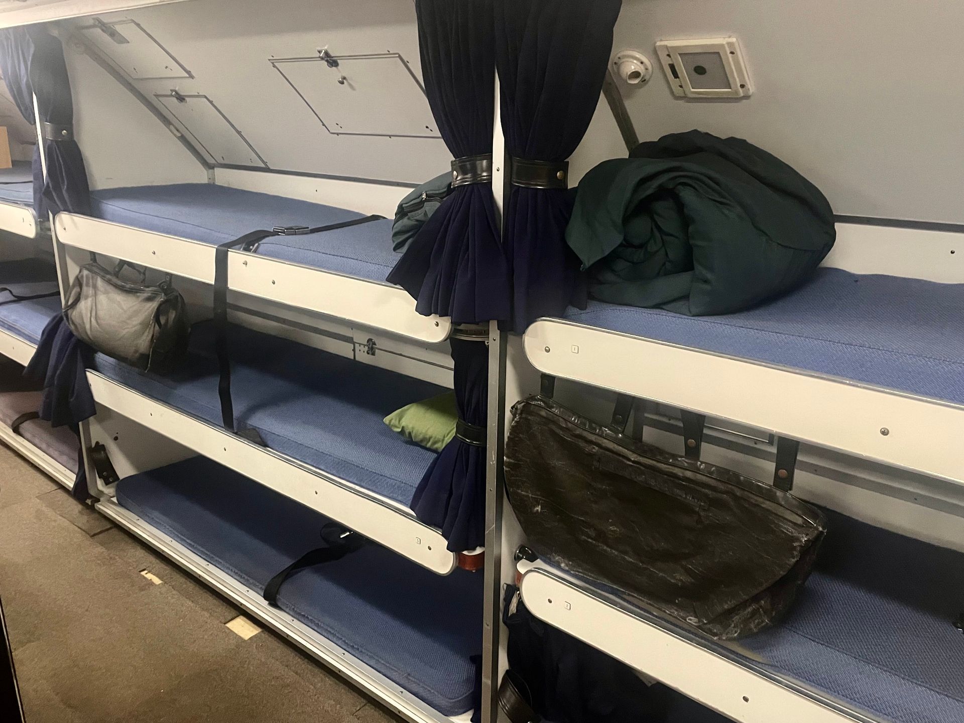 A row of cramped bunks, stacked three high with thin blue mattresses. There are straps like seatbelts across them for securing the crew in rough conditions.