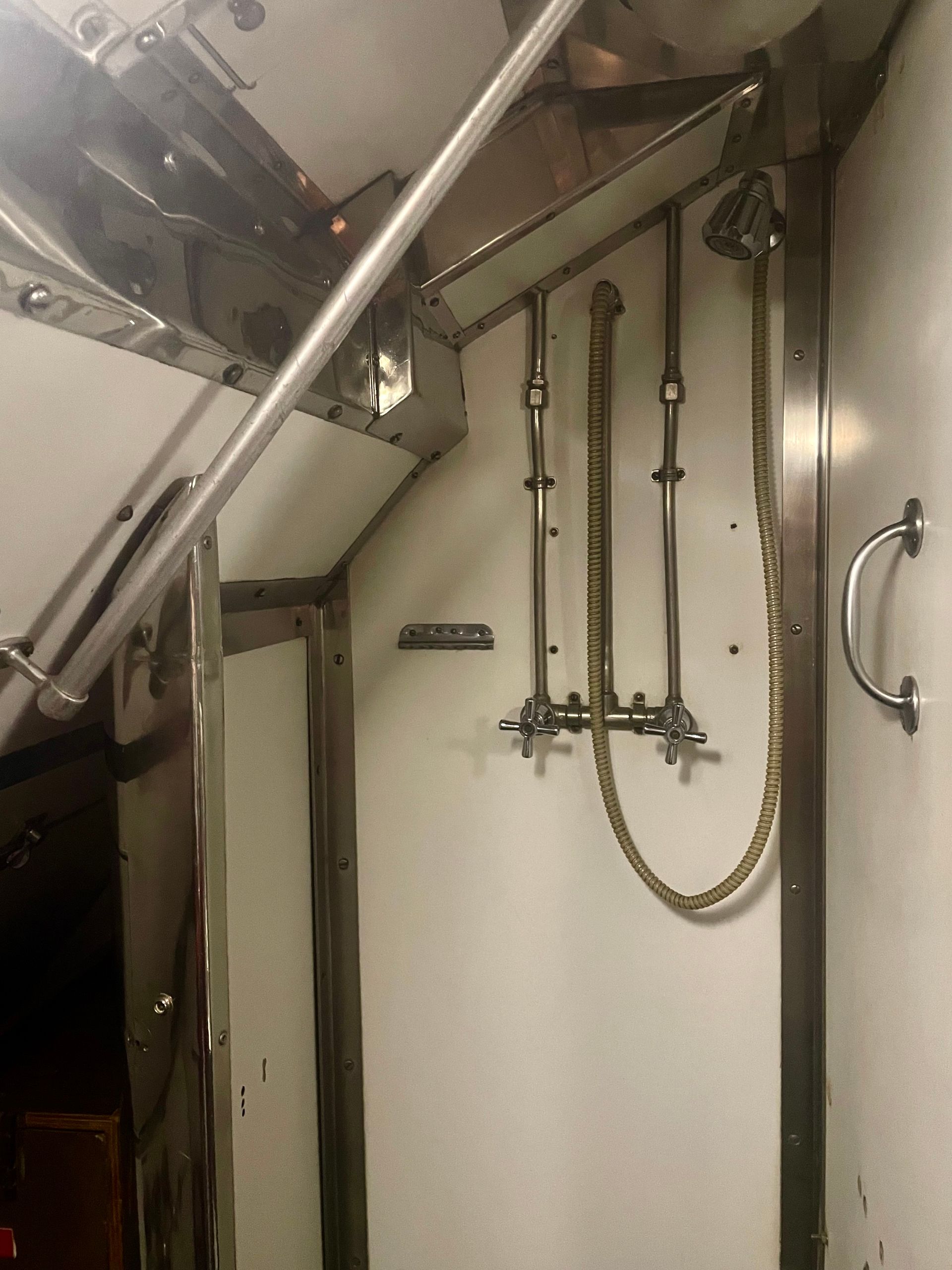 A very bare-bones shower stall with metal taps and shower head on a hose.