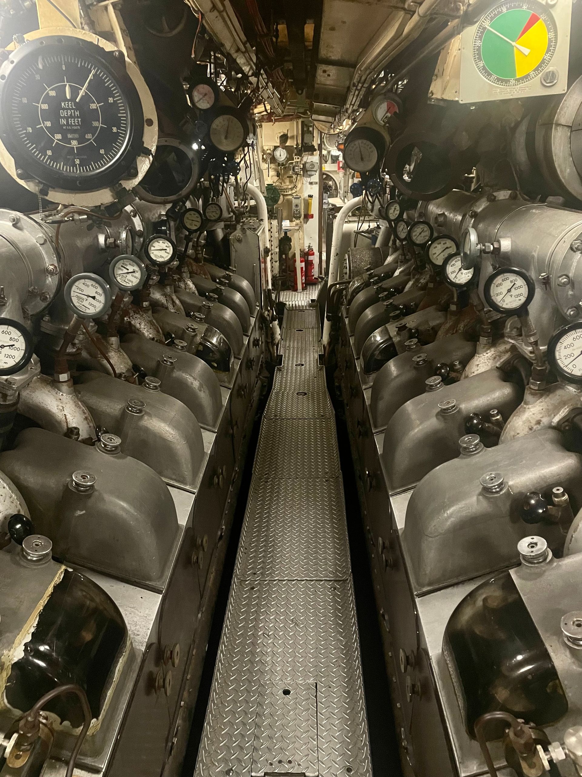 A very narrow walkway between two large engines. Many dials are all over the engines, the closest one reads "Keel depth in feet."