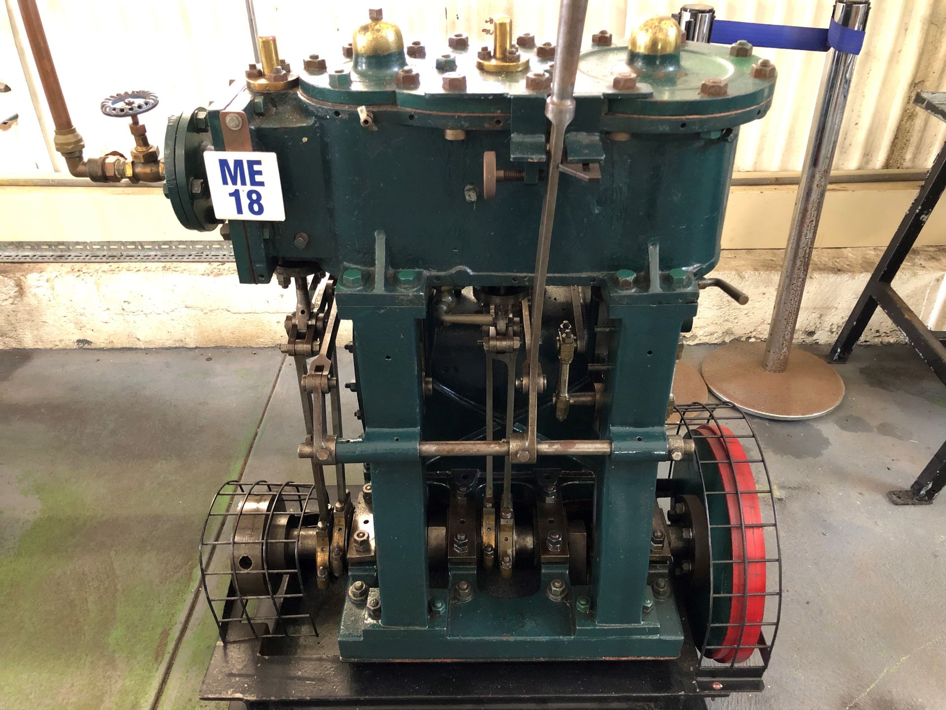 A tall green engine mounted on the floor, a red wheel is on the side at the bottom.