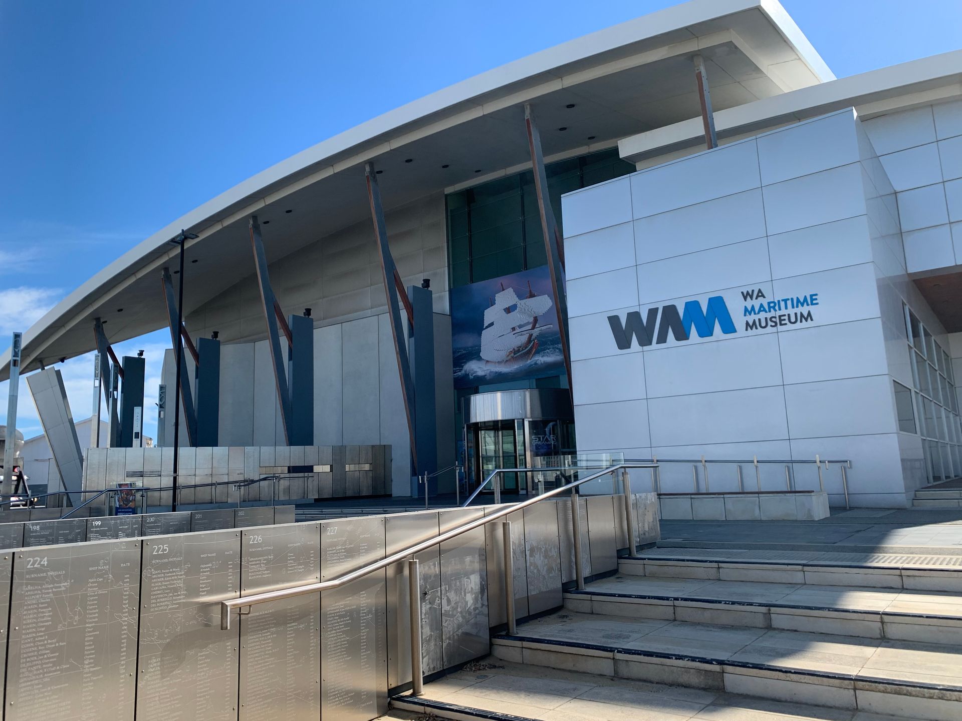 In the foreground are silver panels with writing on them. Steps lead up to a large white and grey building with the name WA Maritime Museum.