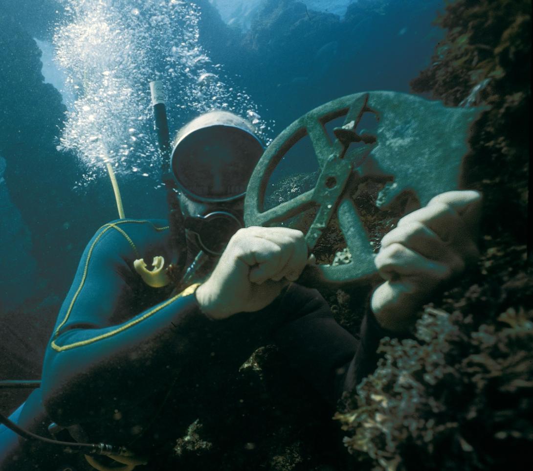 A diver underwater holding up a corroded astrolabe triumphantly.