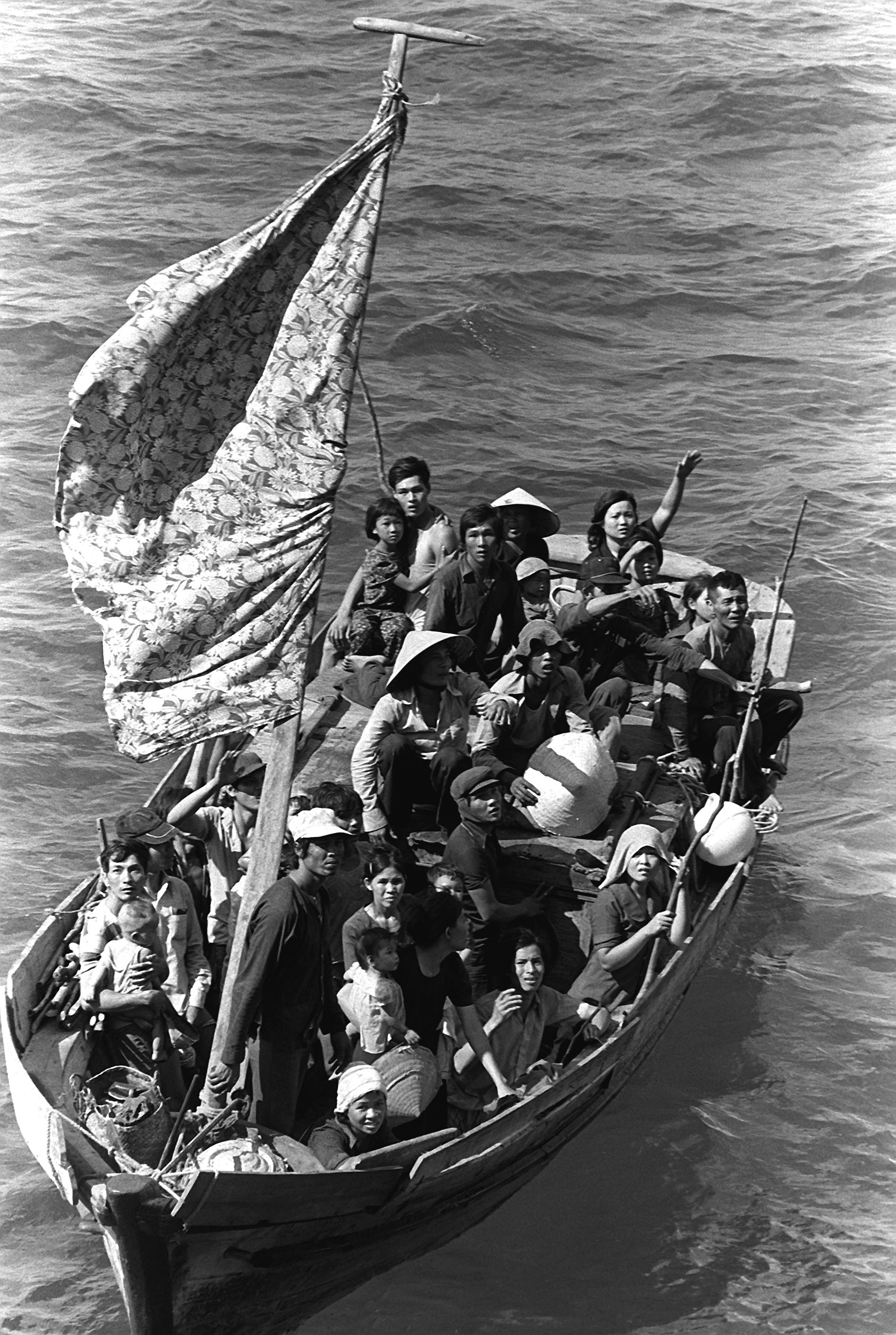Men, women and children crowded on a small wooden boat with makeshift sail. Some are waving for help at a larger ship.
