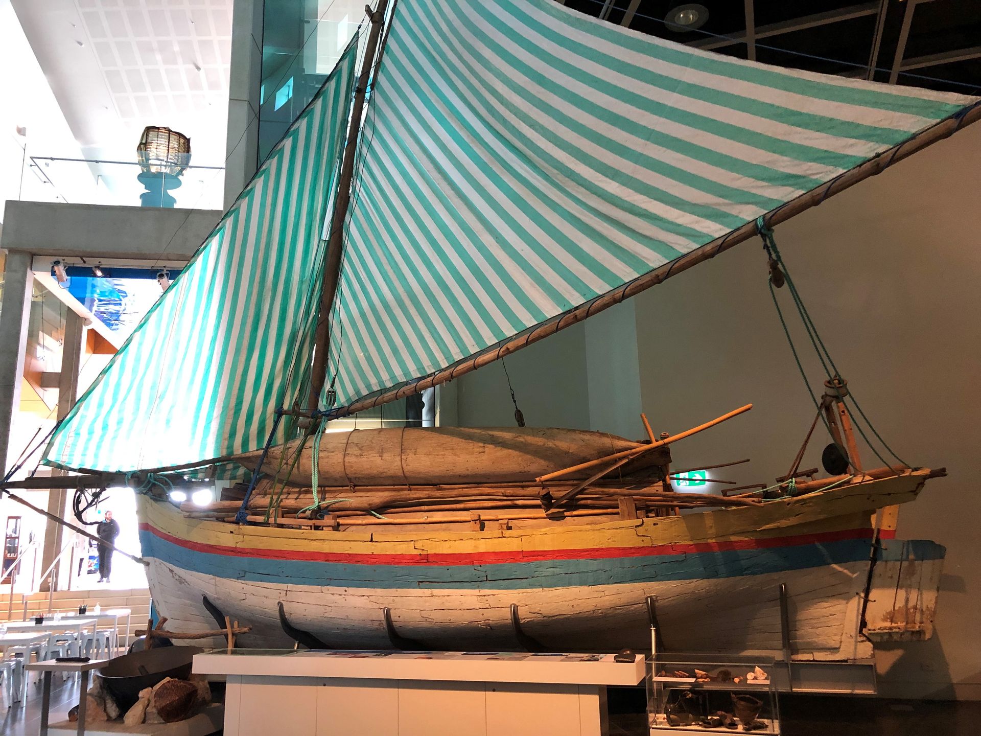 A small wooden sailing boat. The hull is painted white with yellow, red and blue stripes. The sails are striped blue and white.