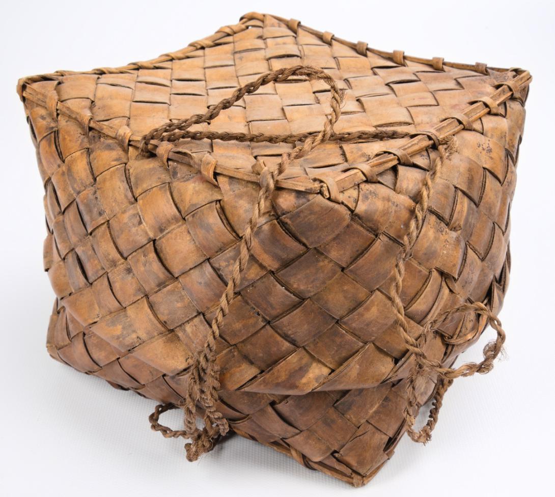The basket is woven from brown material in a diagonal pattern. It is roughly cube shaped and has strings attached for carrying.