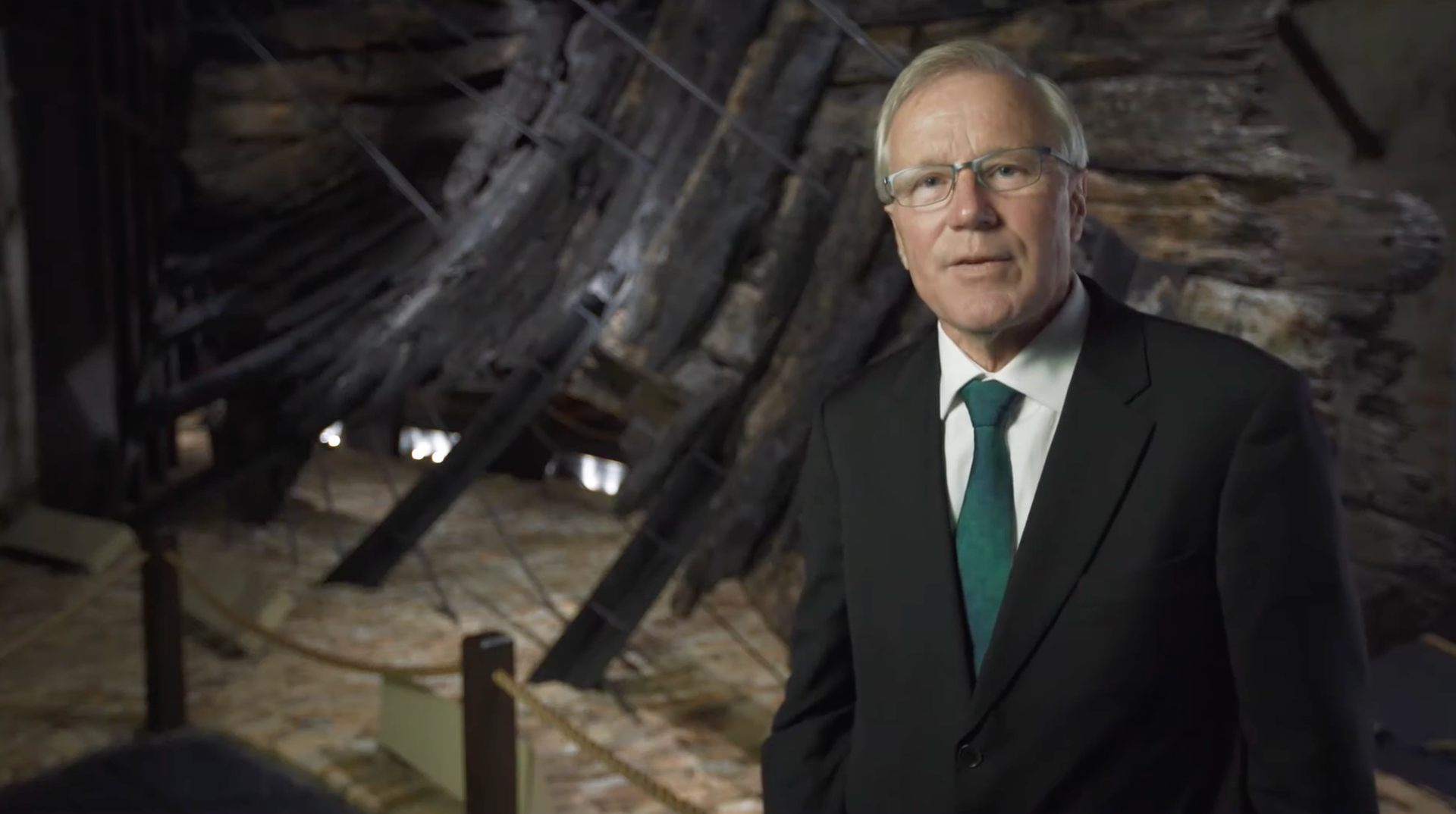 A man in a suit standing in front of the timbers of the Batavia shipwreck.