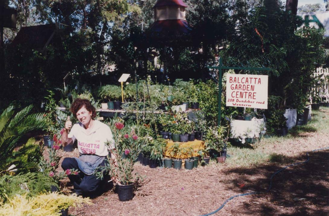 Kneeling at a plant giving a thumbs up, Lena wears a "Balcatta Garden Centre" t-shirt.