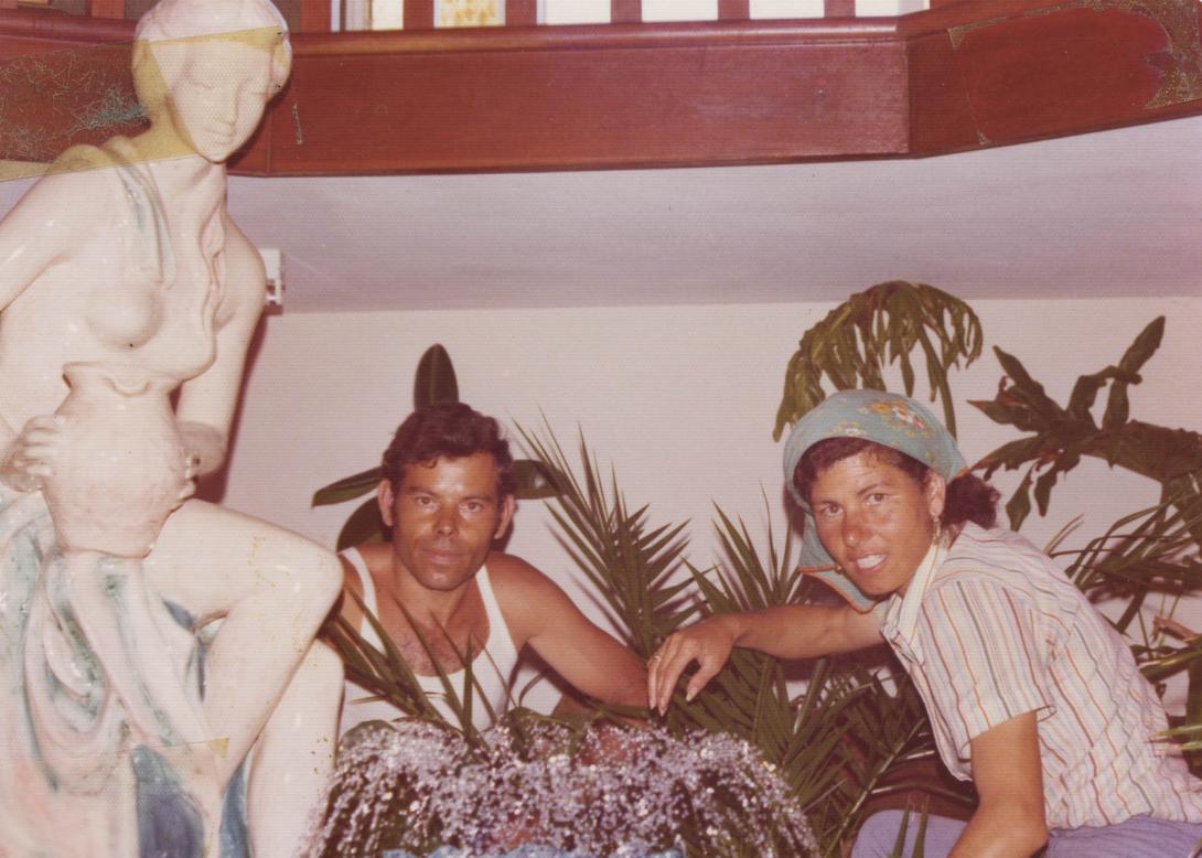 Resting between gardening, a man in a singlet and Lena in a bandanna sit beside a classical statue of a woman with a vase.
