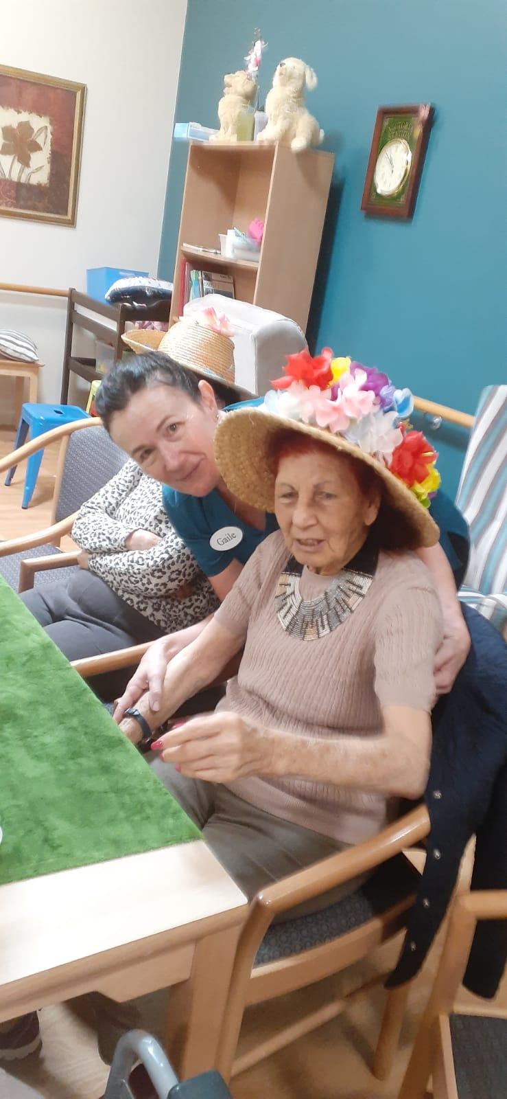 Lady sitting in a residential home wearing a straw hat with brightly coloured flowers.