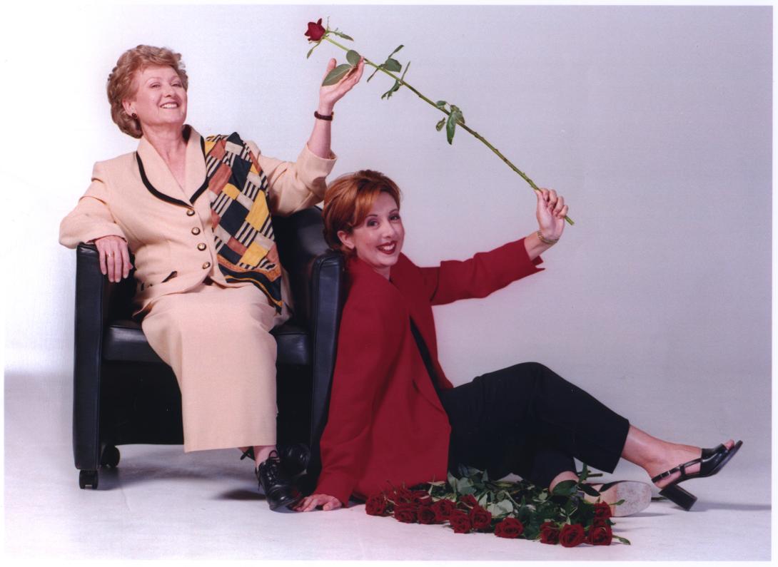 Mother and daughter pictured both holding a rose, with more roses lying on the floor. The mother is sitting on a chair. The daughter is sitting on the phone resting against the chair. They are both smiling.