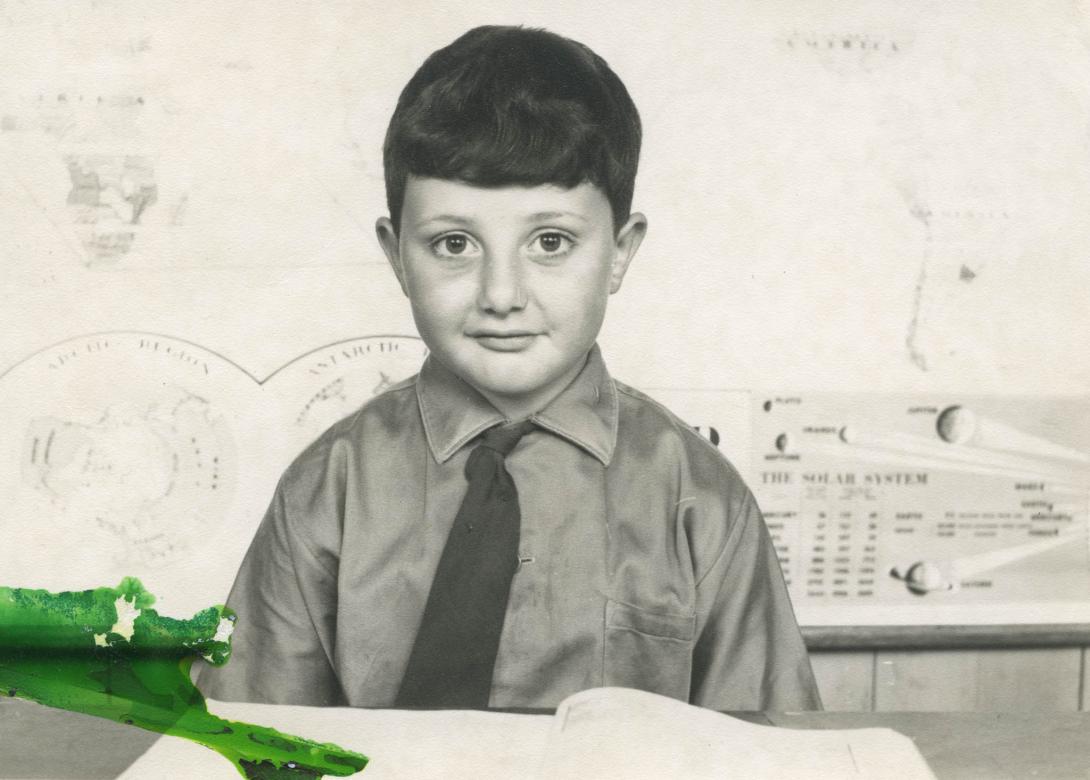 Tony as a school boy in shirt and tie at a desk with school work.