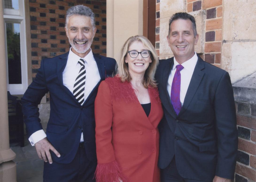 The three parliamentarians in suits smile in front of a brick building.