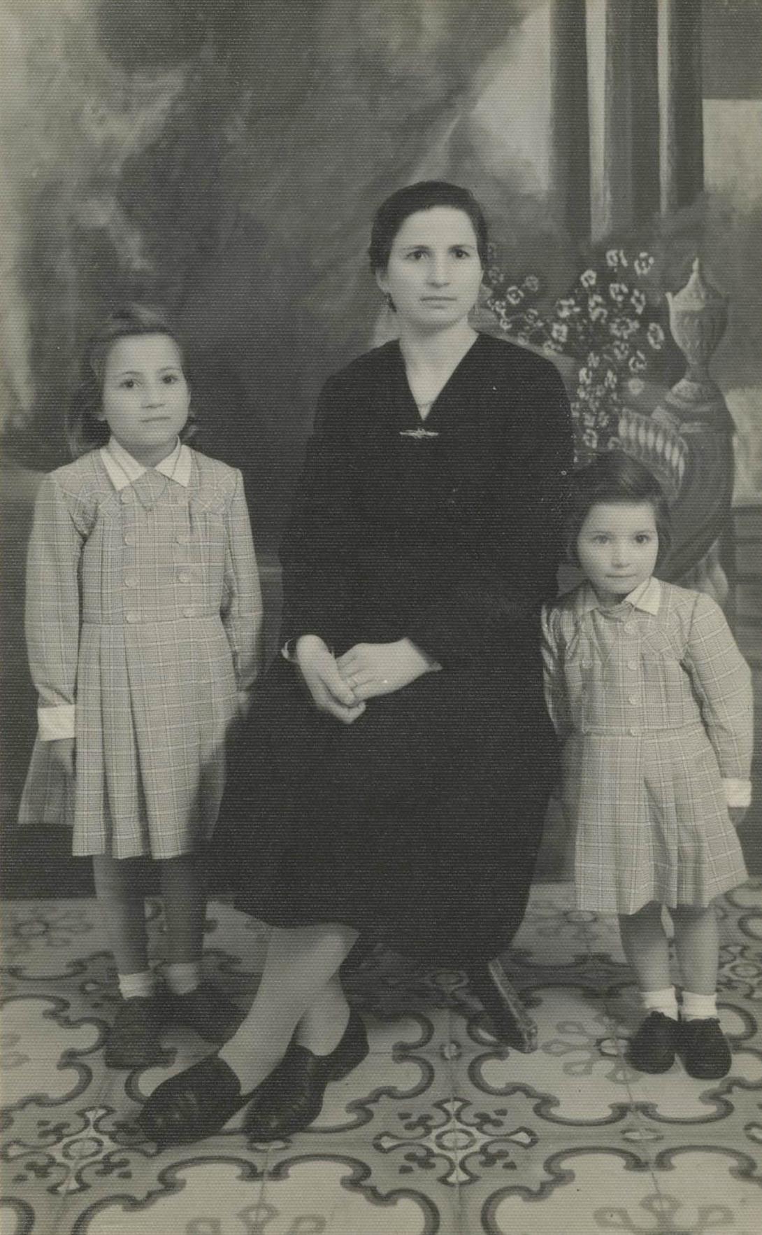 The younger daughter clutches at the mother's dress as they pose formally in a studio.