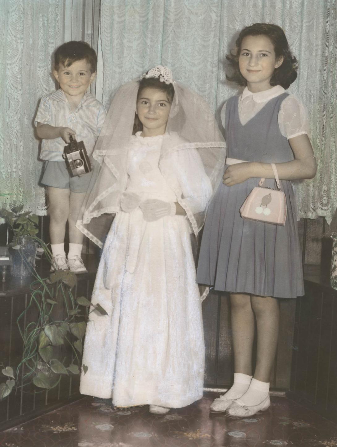 A grinning boy holds up an old box camera with a young girl in a long white dress and veil, and another in a short dress.