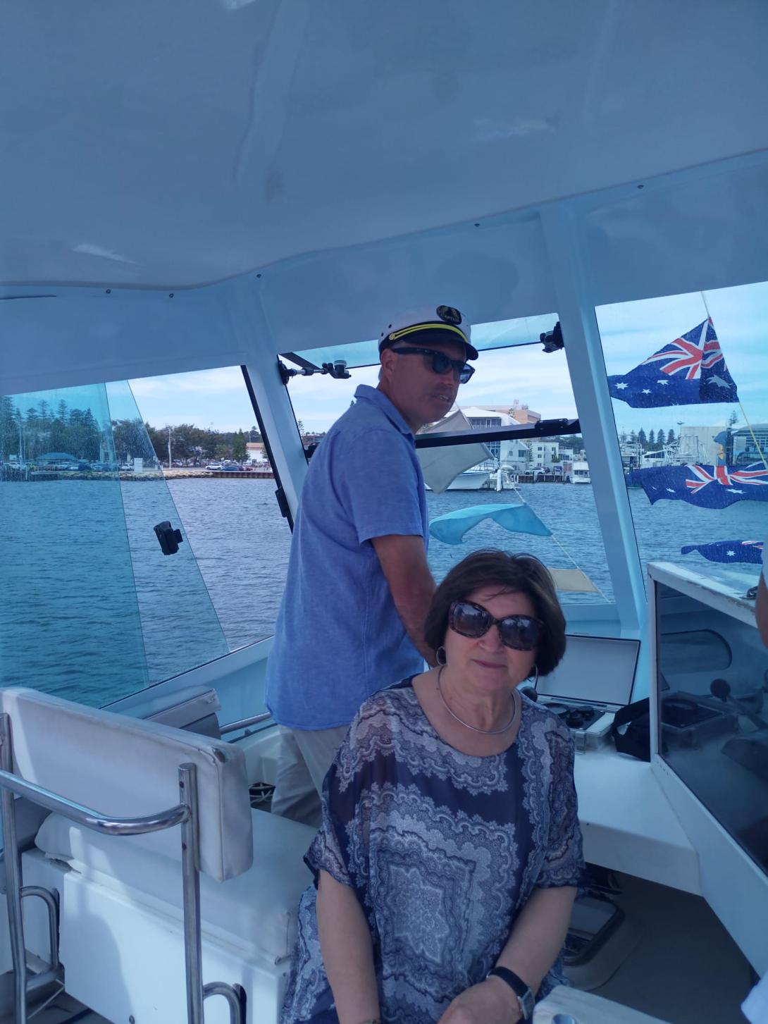 Anna turns to the camera in the boat's cabin as the man steers, wearing a captain's cap.