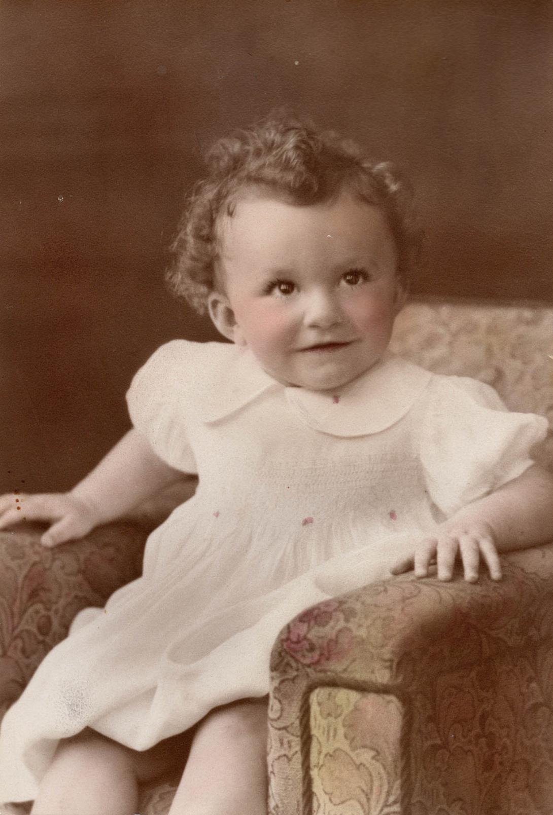 A colourised black & white photo of a child in a white dress smiling at someone to the side of the camera.