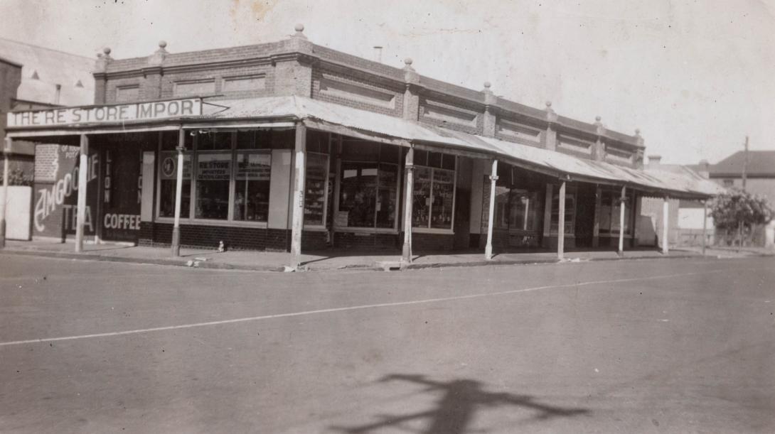 Across a wide empty street, the store advertises Amgoorie Tea and Lion Coffee.