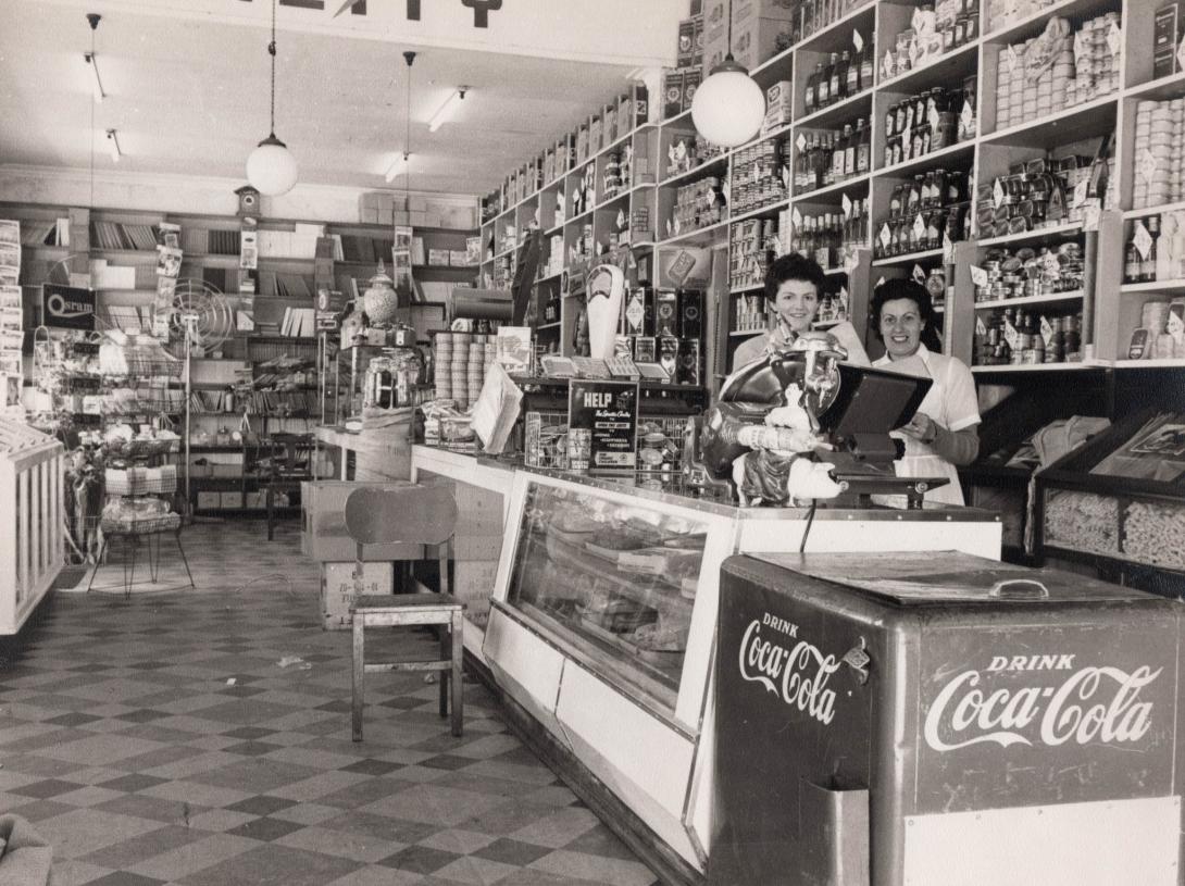 The women smile from behind a deli meat slicer and a cooler advertising "Drink Coca-Cola."