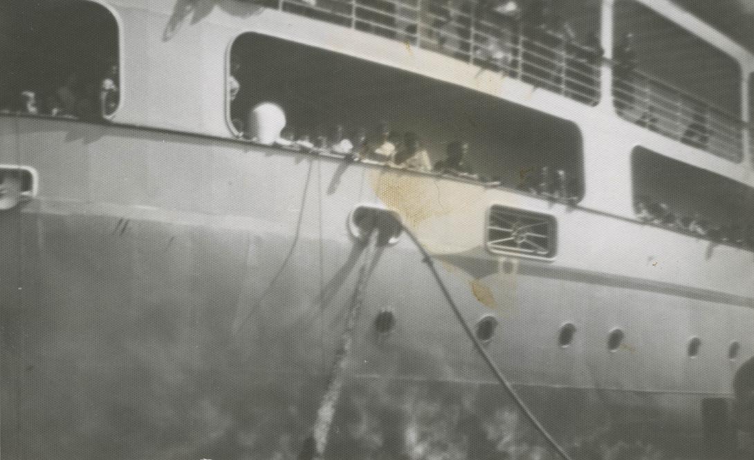 Grainy photo of the side of a large passenger ship. Passengers can be seen on deck looking out for relatives.