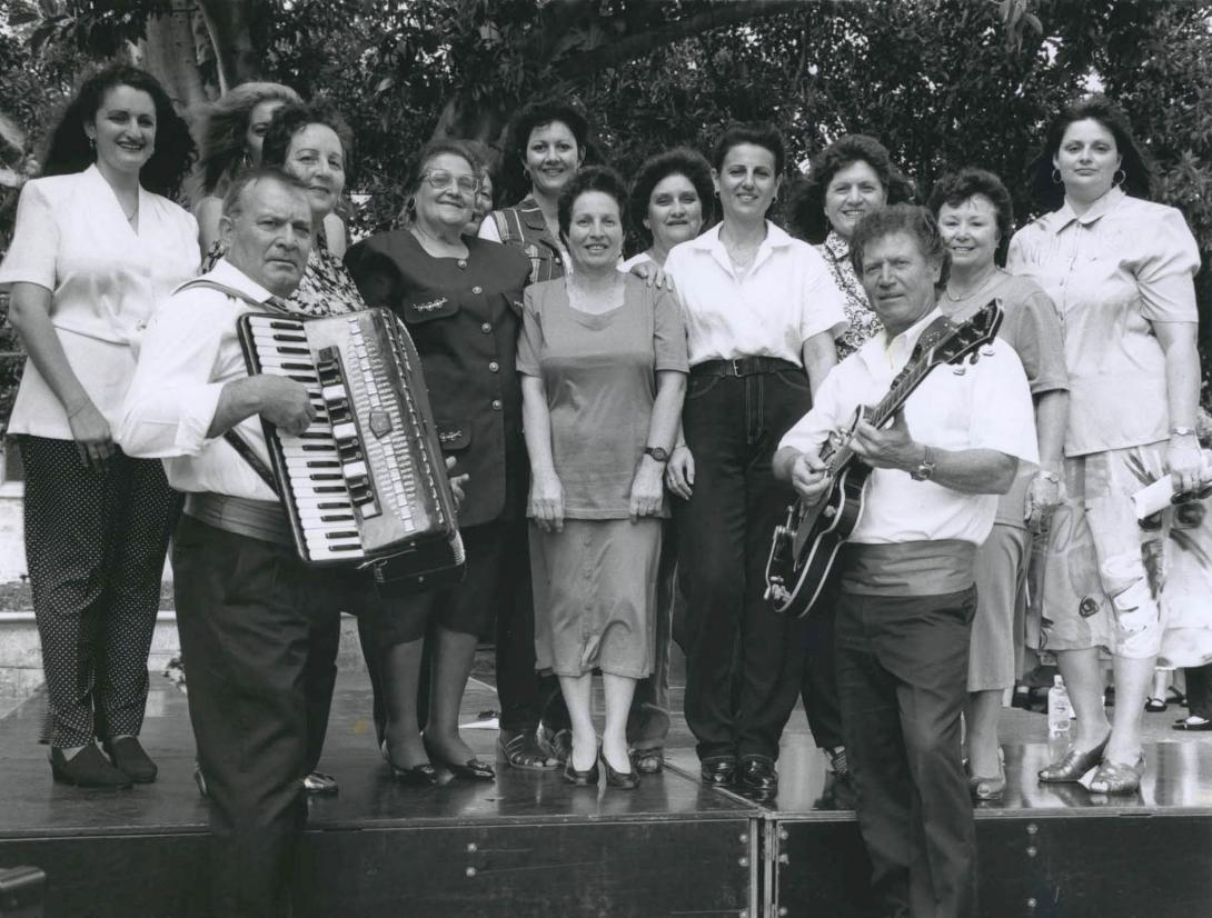 The choir stands on stage, two men with a piano accordion and guitar accompanying.