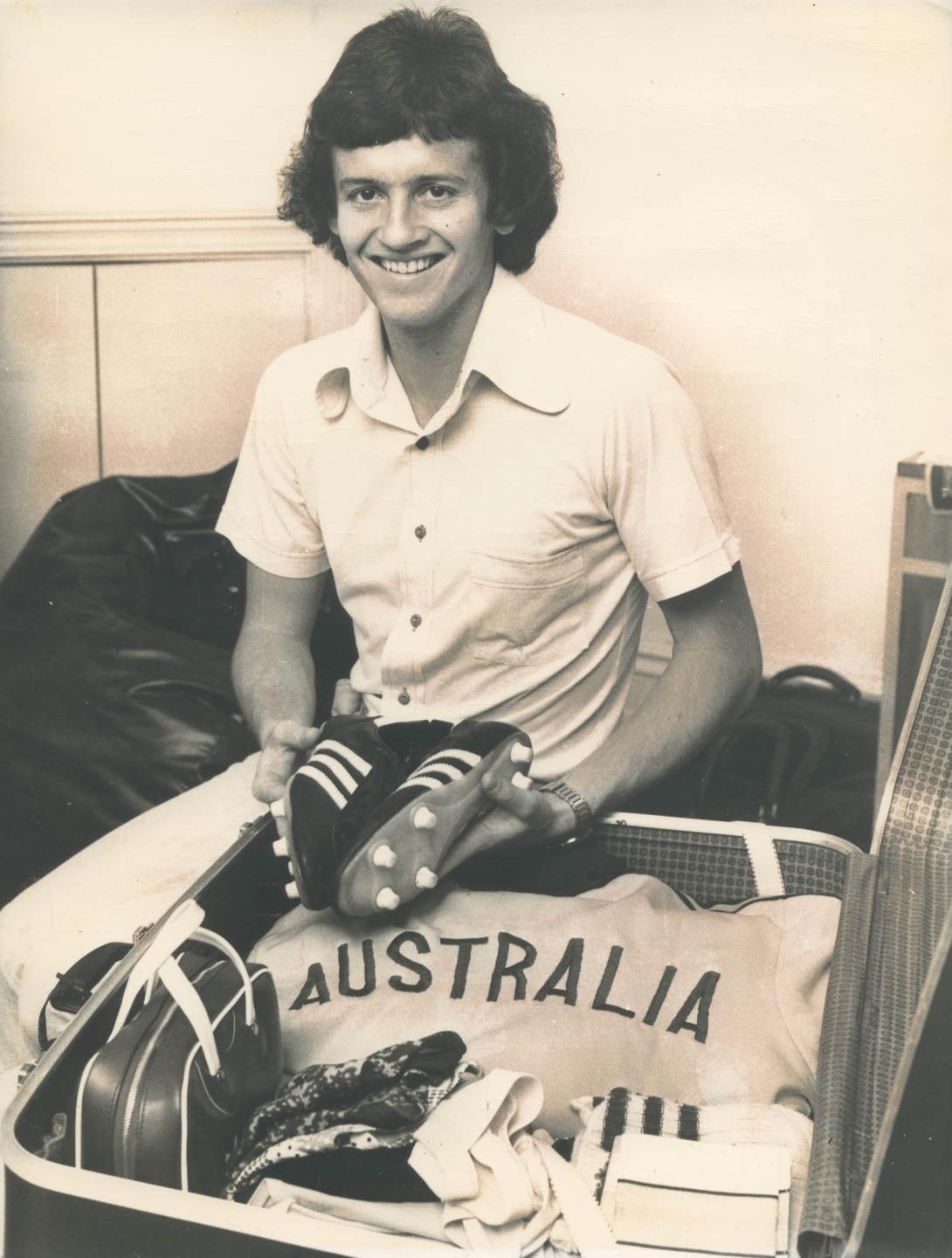 Gary in a locker room holding football shoes and a big bag marked "Australia."