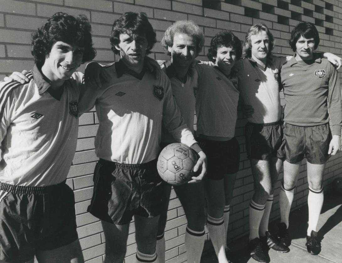 A group of soccer players against a wall, their arms over each others' shoulders.