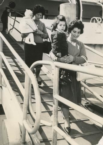 A smiling Vittoria and Maria carrying their children down a passenger ship's gangway.