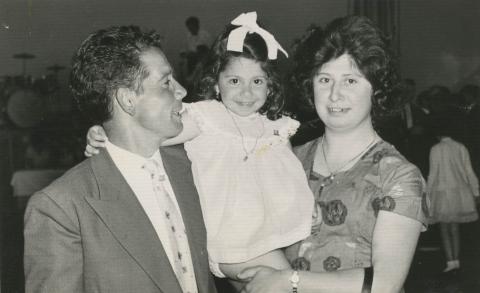 Claudia beams at the camera between her parents, who are holding her up.