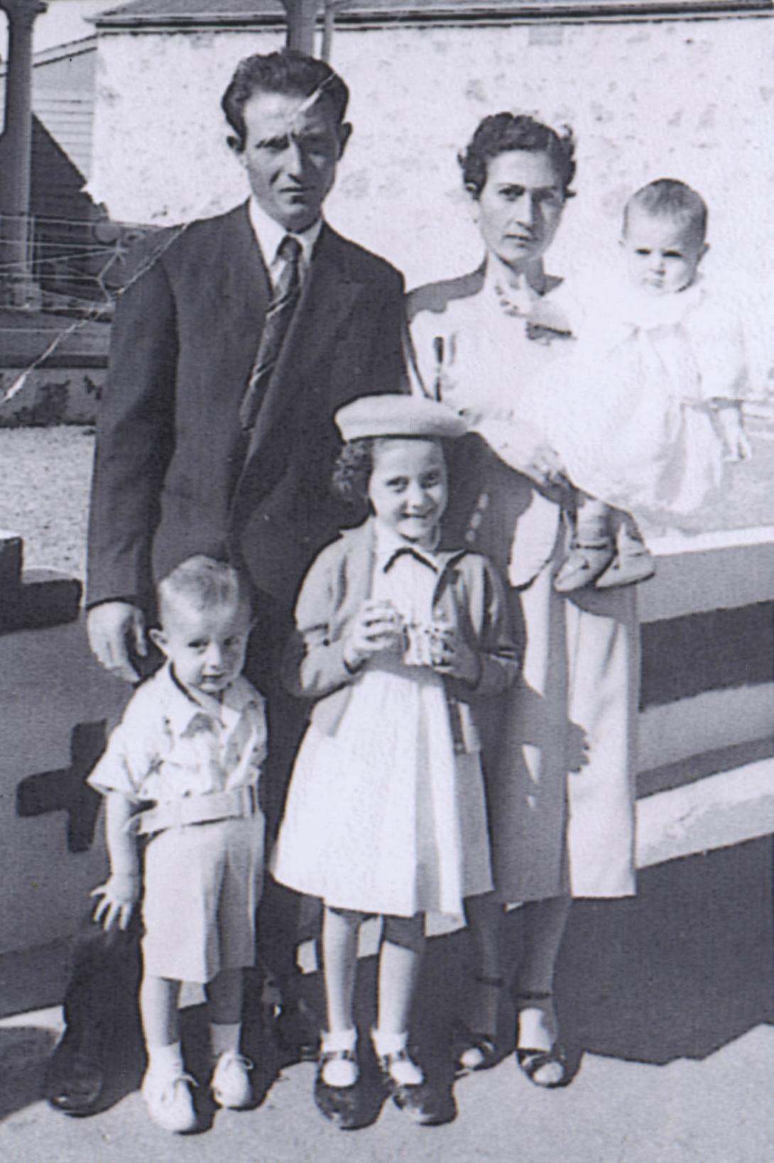 The oldest child, in a dress and beret, smiles between her parents. The younger child and the baby seem less enthusiastic.