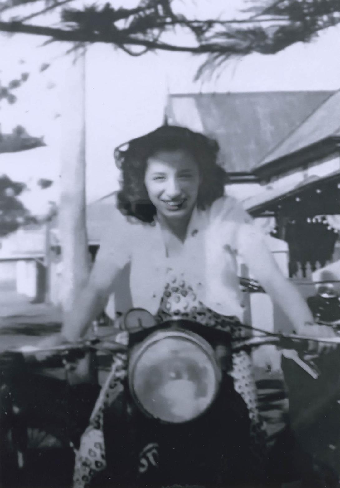 Frances grins on the bike in a dress and short-sleeved jacket, appearing to race right at the camera.